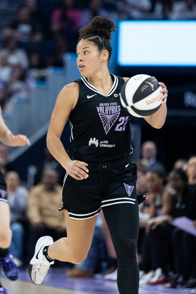 A basketball player in a black "Golden State Valkyries" uniform holds a ball while moving, focused and poised. The background shows blurred spectators.